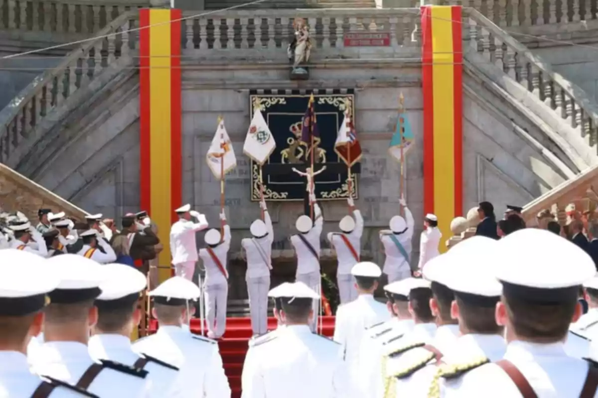 Military ceremony with uniformed officers carrying flags in front of a staircase decorated with the colors of the Spanish flag and a coat of arms in the center Military ceremony with uniformed officers carrying flags in front of a staircase decorated with the colors of the Spanish flag and a coat of arms in the center