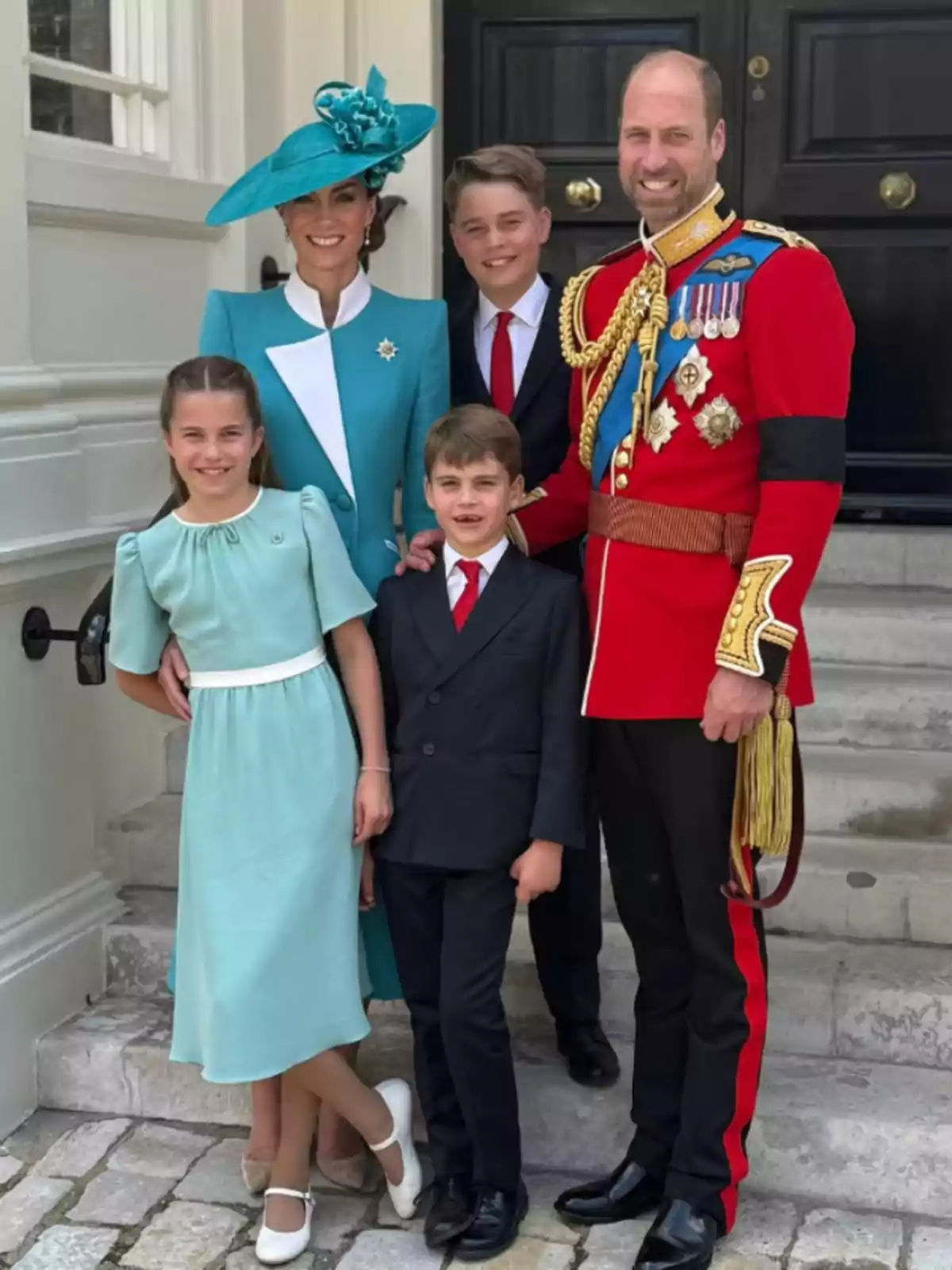 Una familia elegantemente vestida posa sonriente en la entrada de un edificio, con el hombre usando un uniforme rojo con medallas y la mujer un vestido azul con sombrero a juego, acompañados de tres niños bien arreglados. Una familia elegantemente vestida posa sonriente en la entrada de un edificio, con el hombre usando un uniforme rojo con medallas y la mujer un vestido azul con sombrero a juego, acompañados de tres niños bien arreglados.