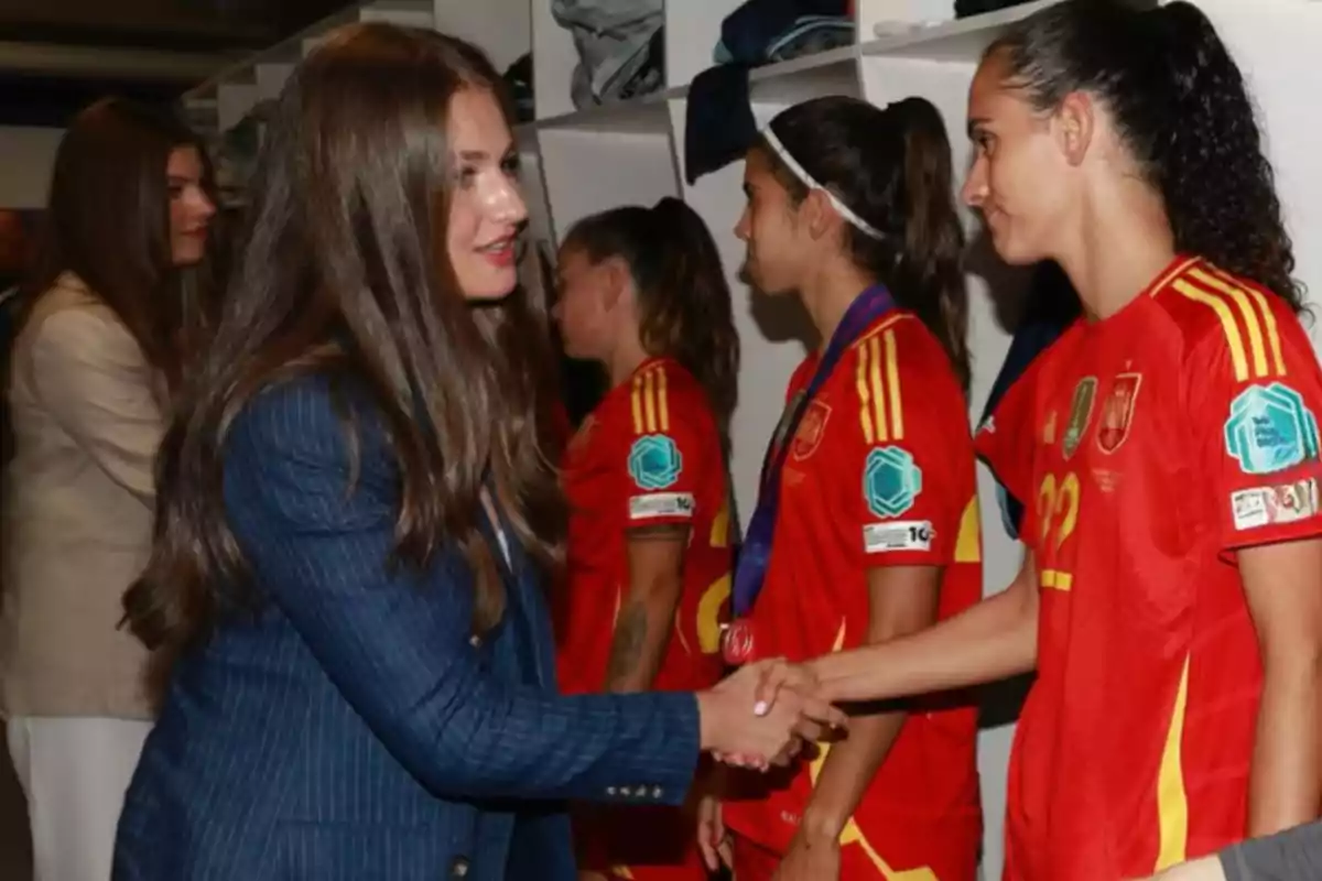 A woman in a blue suit greets a player wearing the red uniform of the Spanish national team inside a locker room. A woman in a blue suit greets a player wearing the red uniform of the Spanish national team inside a locker room.