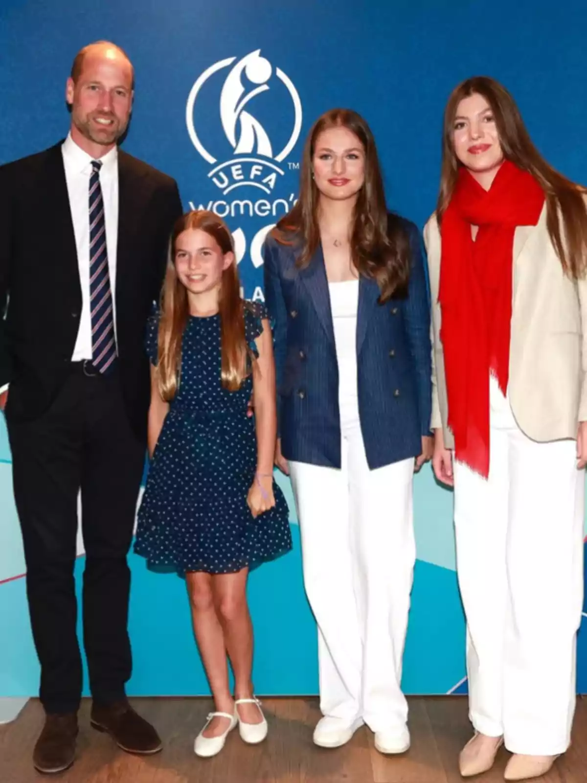Four people pose smiling in front of a blue background with the UEFA Women’s Euro logo, two adults and two girls, all dressed elegantly and casually. Four people pose smiling in front of a blue background with the UEFA Women’s Euro logo, two adults and two girls, all dressed elegantly and casually.