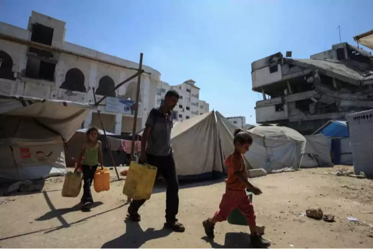A family walks among tents in an improvised camp carrying water jugs, with damaged buildings in the background under a clear sky. A family walks among tents in an improvised camp carrying water jugs, with damaged buildings in the background under a clear sky.