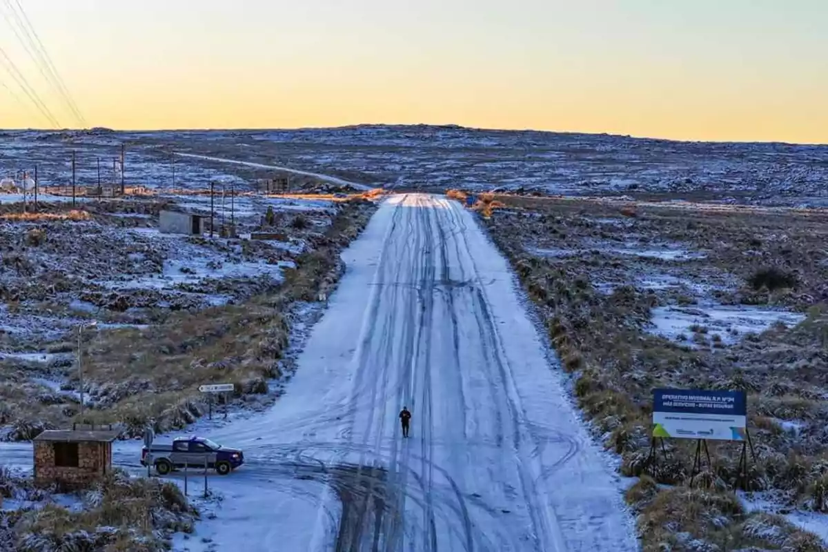 Carretera cubierta de nieve al amanecer con una persona caminando en el centro y una camioneta estacionada a la izquierda