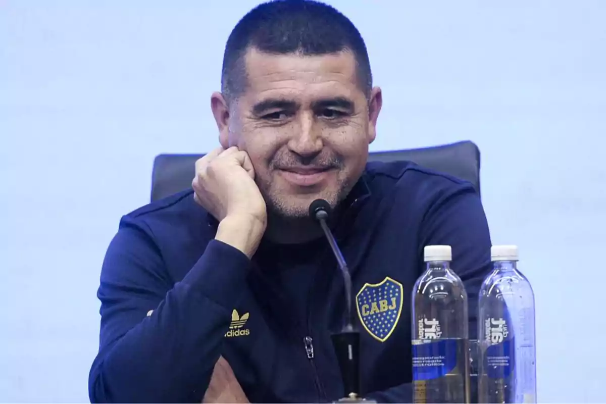 Hombre con chaqueta azul de Boca Juniors sonriendo en una conferencia de prensa con dos botellas de agua frente a él