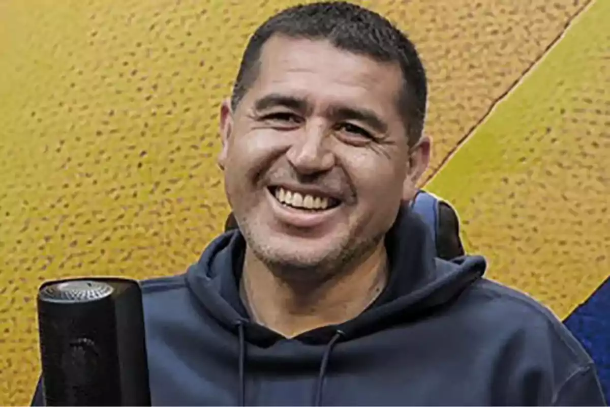 Hombre de cabello corto y oscuro sonríe frente a un fondo amarillo mientras usa una sudadera azul y está sentado junto a un micrófono