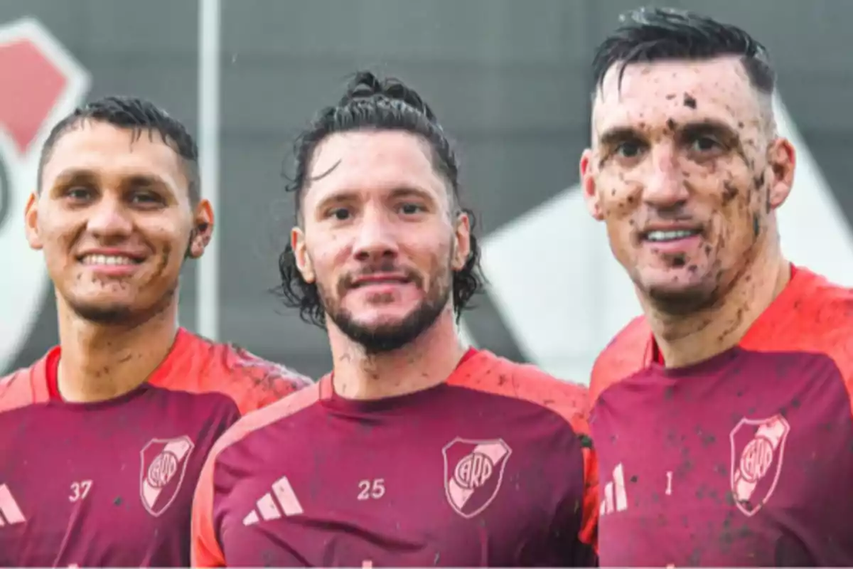 Tres futbolistas con camisetas de entrenamiento de River Plate sonríen a la cámara con manchas de barro en la cara y la ropa. Tres futbolistas con camisetas de entrenamiento de River Plate sonríen a la cámara con manchas de barro en la cara y la ropa.