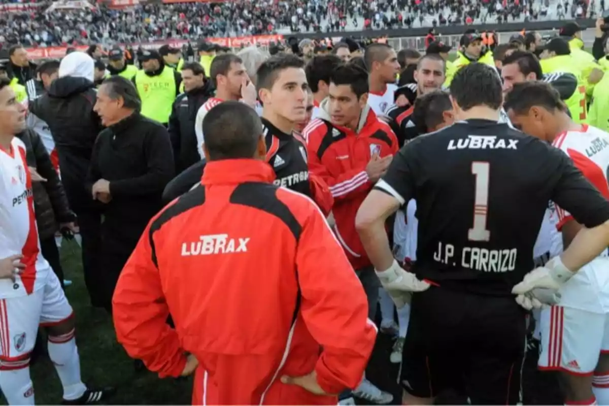 Jugadores y cuerpo técnico de un equipo de fútbol con uniformes blancos y rojos se agrupan en el campo mientras la tribuna está llena de espectadores Jugadores y cuerpo técnico de un equipo de fútbol con uniformes blancos y rojos se agrupan en el campo mientras la tribuna está llena de espectadores