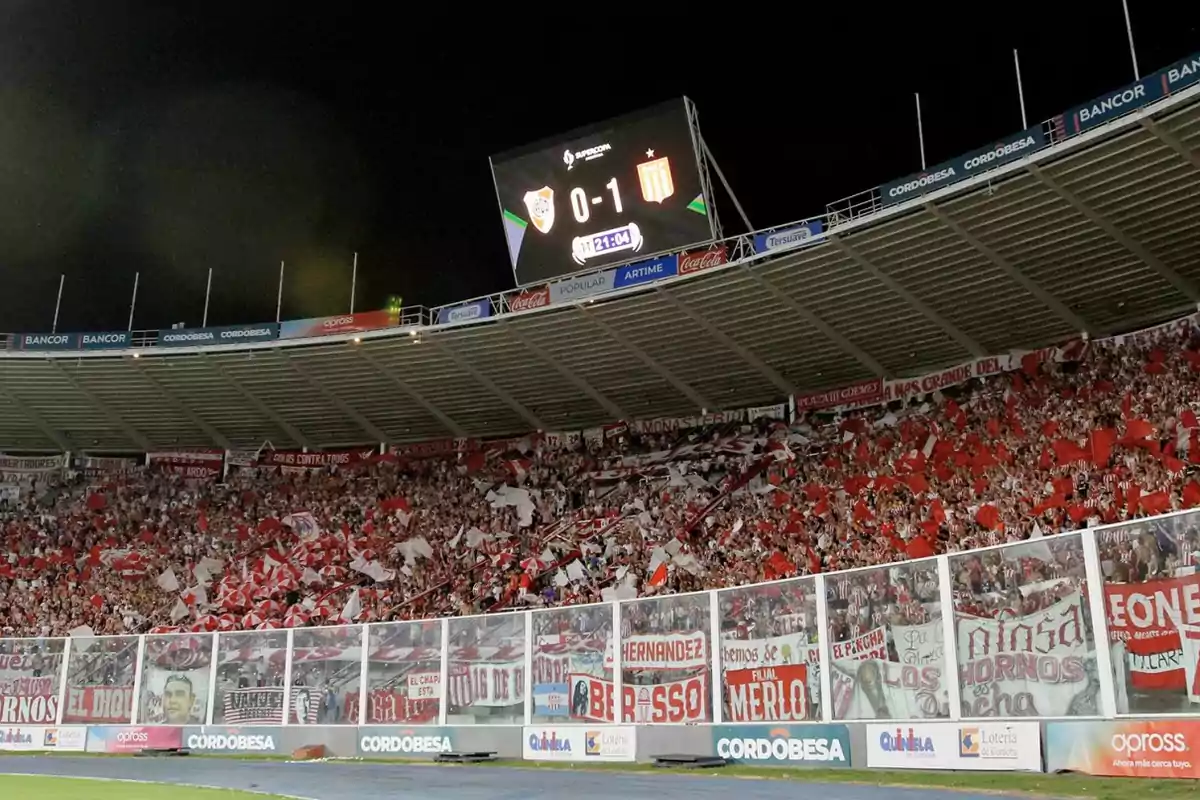 Soccer fans fill a stand with red flags and jerseys while a scoreboard displays the result of a nighttime match. Soccer fans fill a stand with red flags and jerseys while a scoreboard displays the result of a nighttime match.