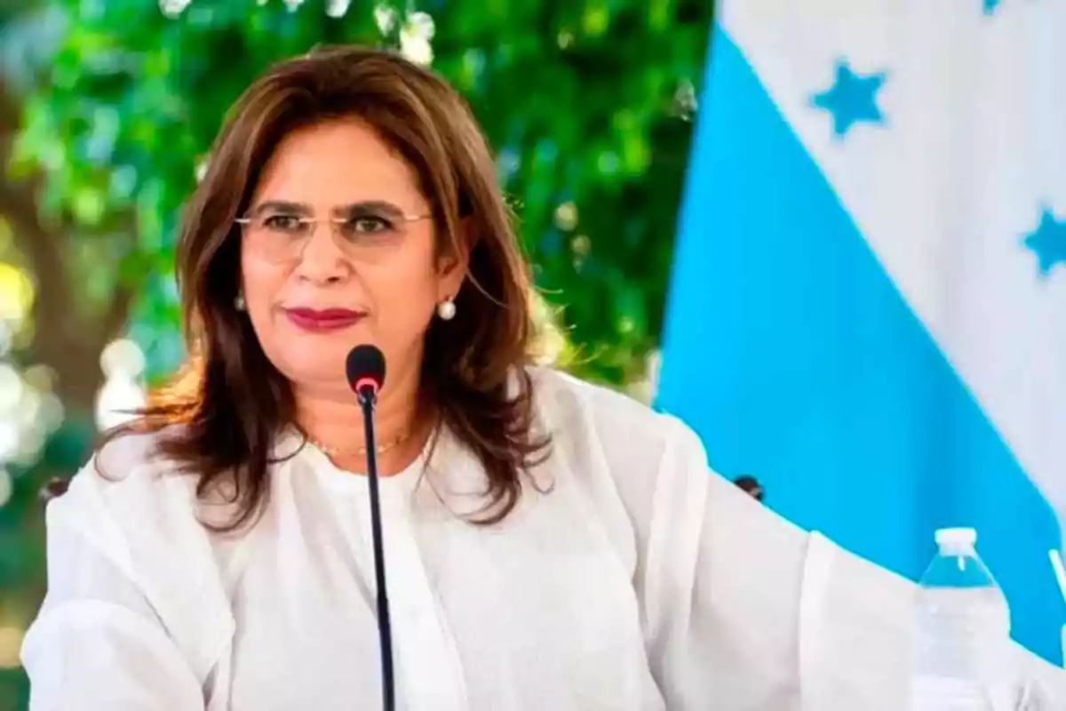 Woman with glasses and a white blouse speaking in front of a microphone with a blue and white flag in the background Woman with glasses and a white blouse speaking in front of a microphone with a blue and white flag in the background