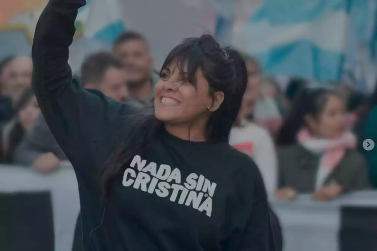 Mujer sonriente con cabello oscuro y flequillo levantando el brazo, lleva una sudadera negra con la frase "Nada sin Cristina" en letras blancas, al fondo se ven personas y banderas argentinas desenfocadas.