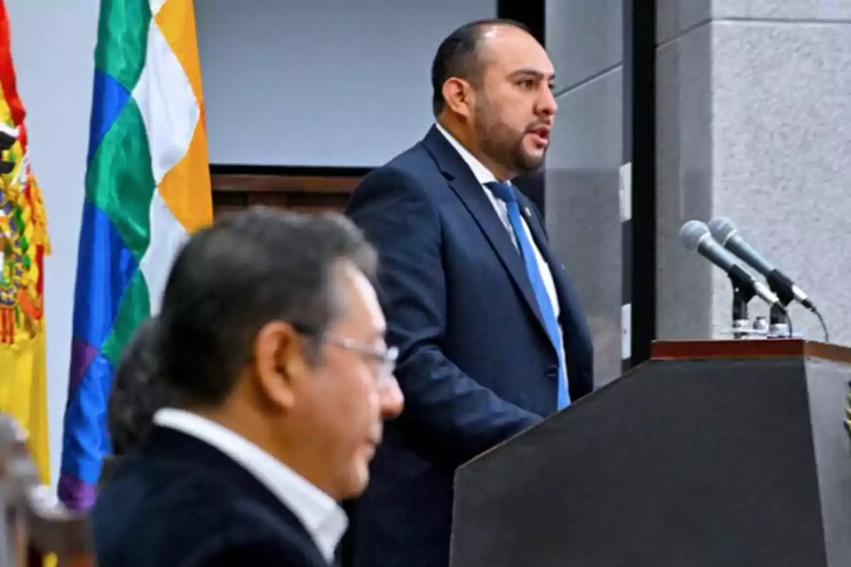 Man in a suit speaking at a podium with microphones while another person is seated in the foreground and flags can be seen in the background Man in a suit speaking at a podium with microphones while another person is seated in the foreground and flags can be seen in the background