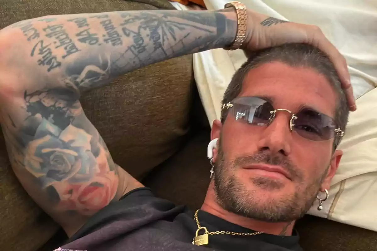 Man lying on a couch wearing sunglasses, with tattoos on his arm, a gold chain, and white headphones