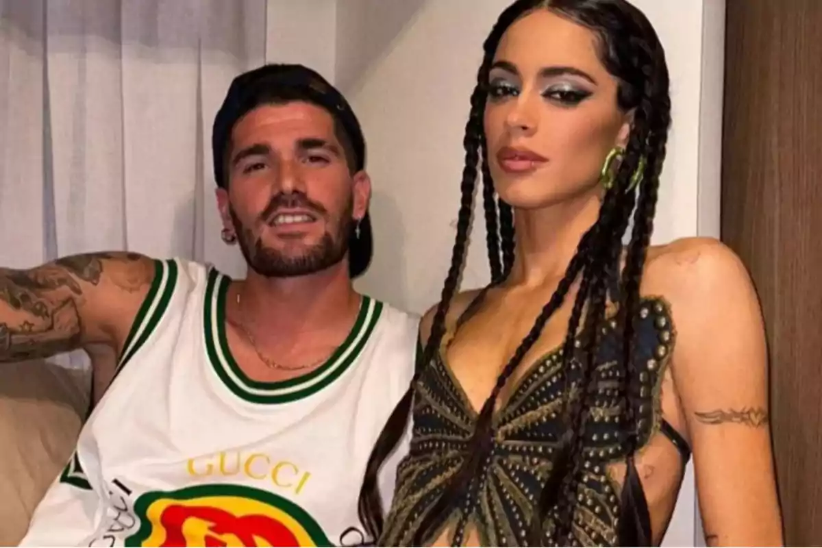 A man in a white tank top and a woman with long braids and a decorated top pose together indoors. A man in a white tank top and a woman with long braids and a decorated top pose together indoors.