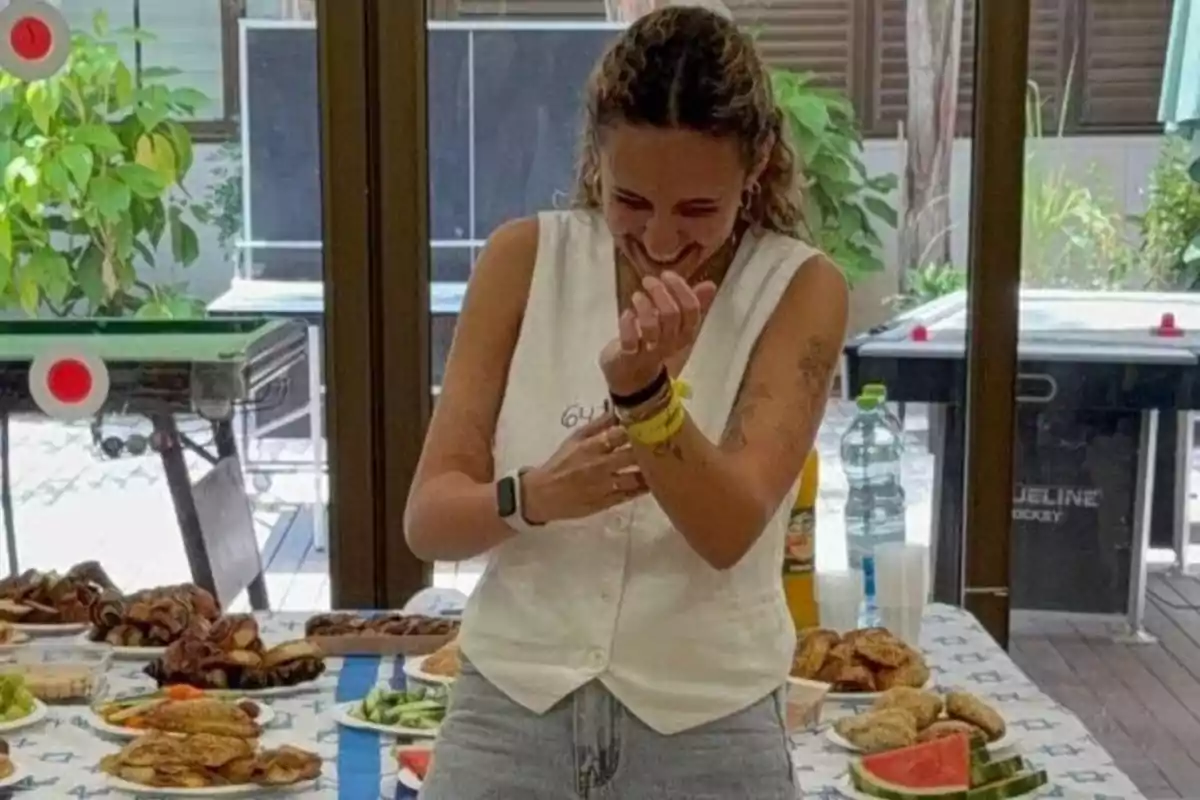 Woman smiling in front of a table with assorted food in an indoor setting with windows and plants in the background