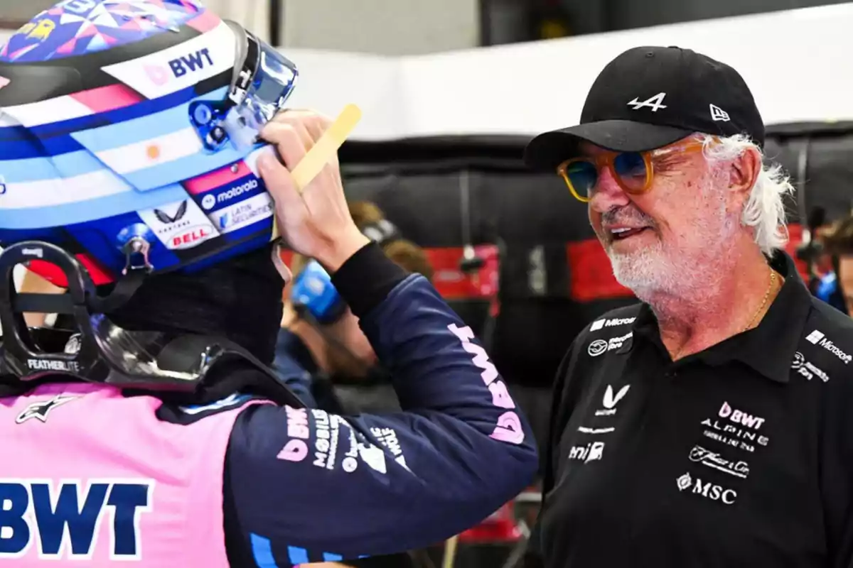 A pilot wearing a helmet and BWT Alpine racing suit smiles as he talks with a man with gray hair, a beard, glasses, and a black cap inside a Formula 1 garage. A pilot wearing a helmet and BWT Alpine racing suit smiles as he talks with a man with gray hair, a beard, glasses, and a black cap inside a Formula 1 garage.