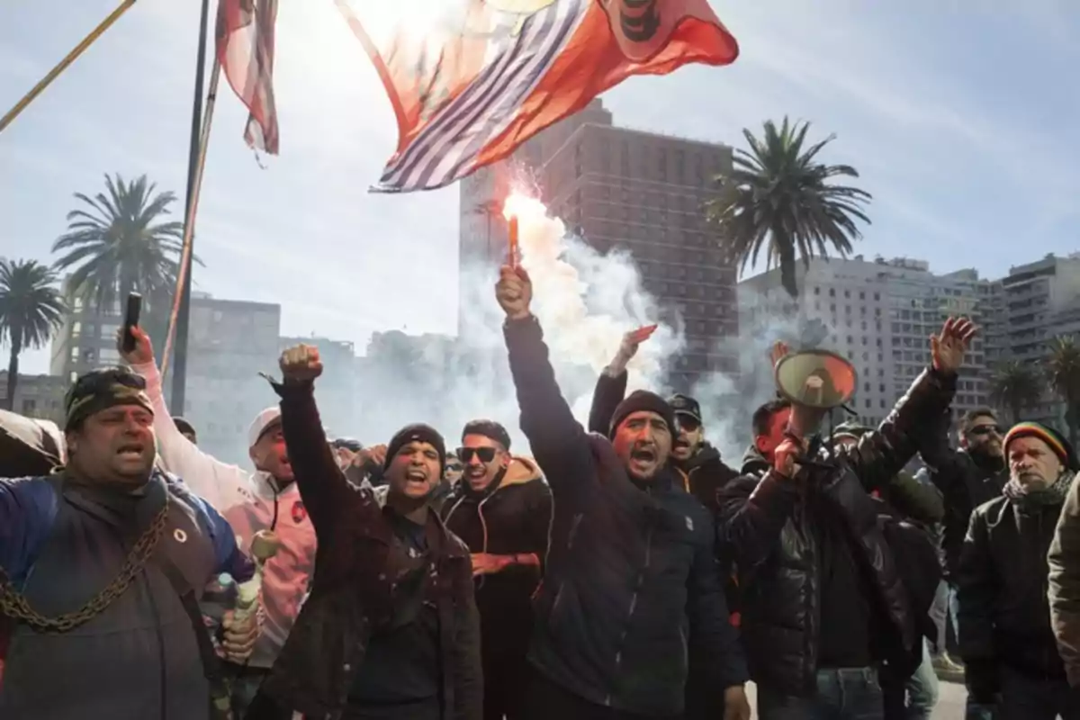Protesters gathered in a square raise their arms and shout; one holds a lit flare and others carry flags, with buildings and palm trees in the background.