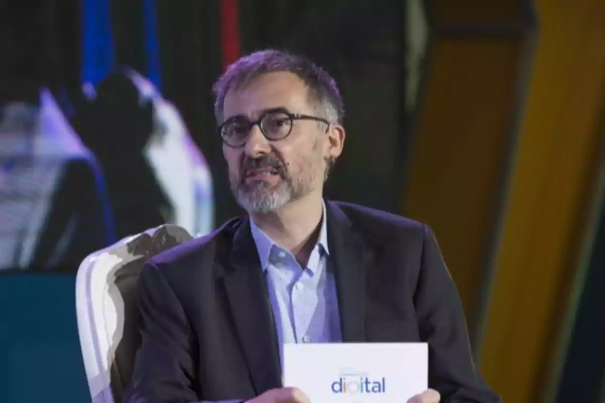 Hombre con gafas y barba sentado en una silla elegante sosteniendo una carpeta blanca con la palabra digital en un escenario iluminado