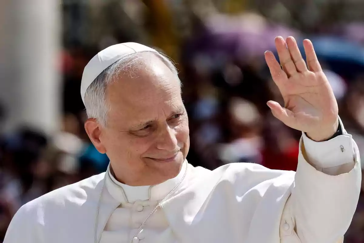 Elderly man dressed in a white cassock and zucchetto waving with his hand raised in front of a blurred crowd