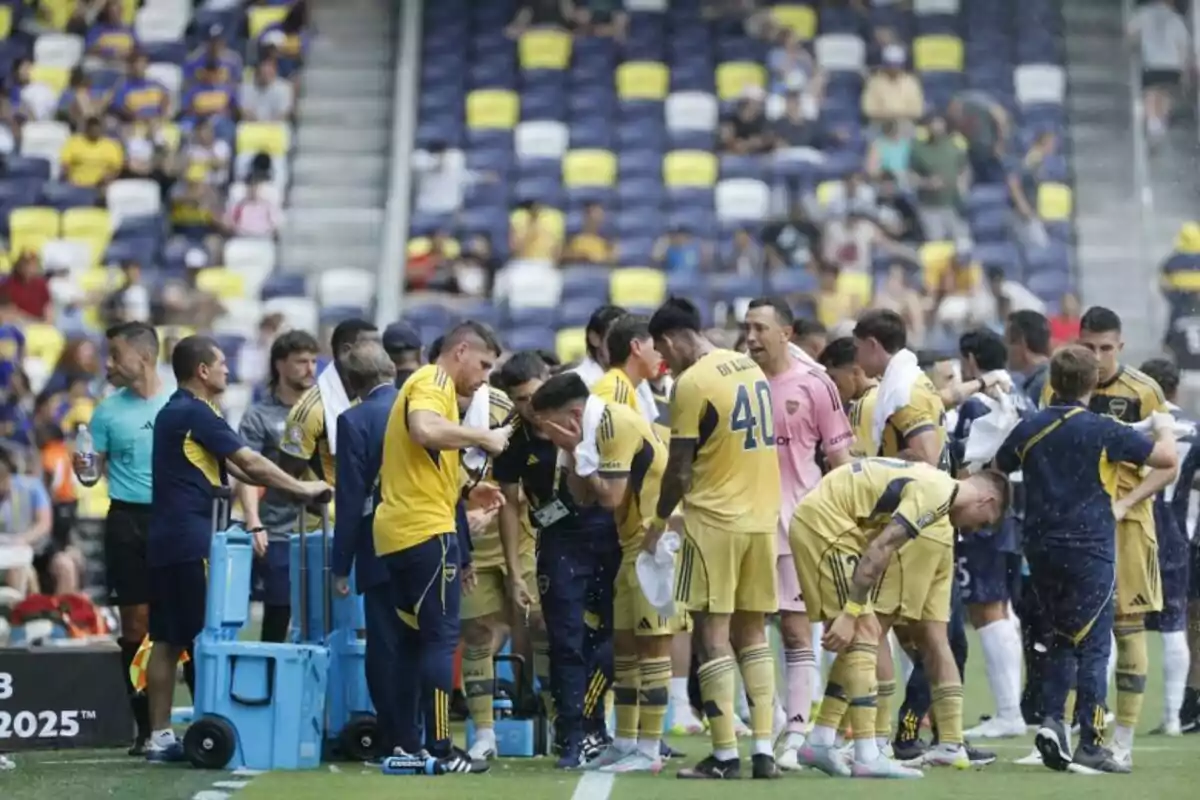Jugadores de fútbol con uniforme amarillo y azul toman un descanso en la banda durante un partido mientras se hidratan y refrescan, con el público en las gradas al fondo. Jugadores de fútbol con uniforme amarillo y azul toman un descanso en la banda durante un partido mientras se hidratan y refrescan, con el público en las gradas al fondo.