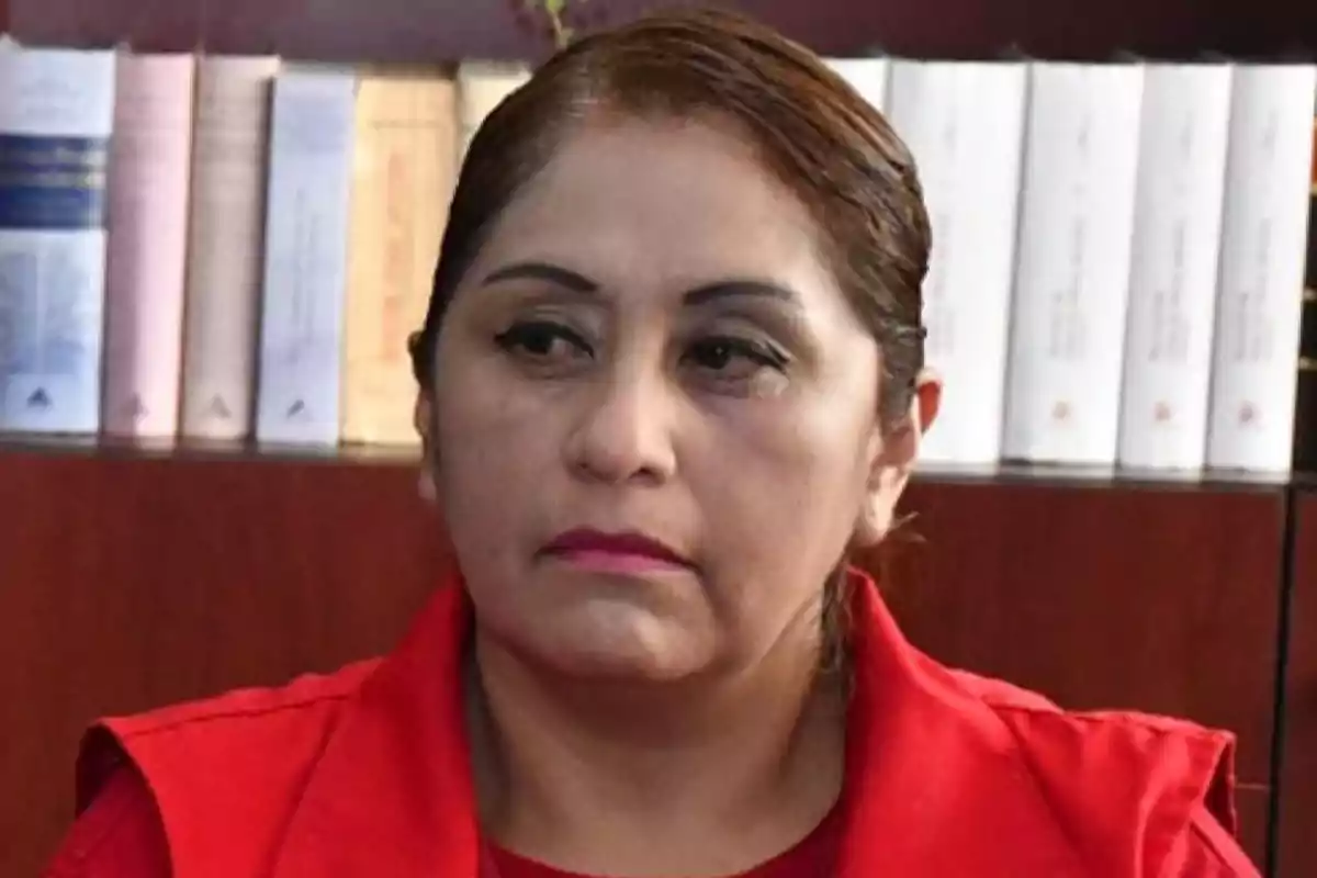 Mujer de cabello recogido y vestimenta roja sentada frente a un estante con libros blancos y beige