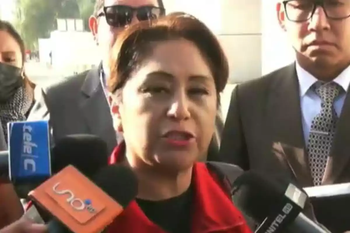 Una mujer con chaqueta roja da declaraciones a la prensa rodeada de micrófonos y personas en traje Una mujer con chaqueta roja da declaraciones a la prensa rodeada de micrófonos y personas en traje