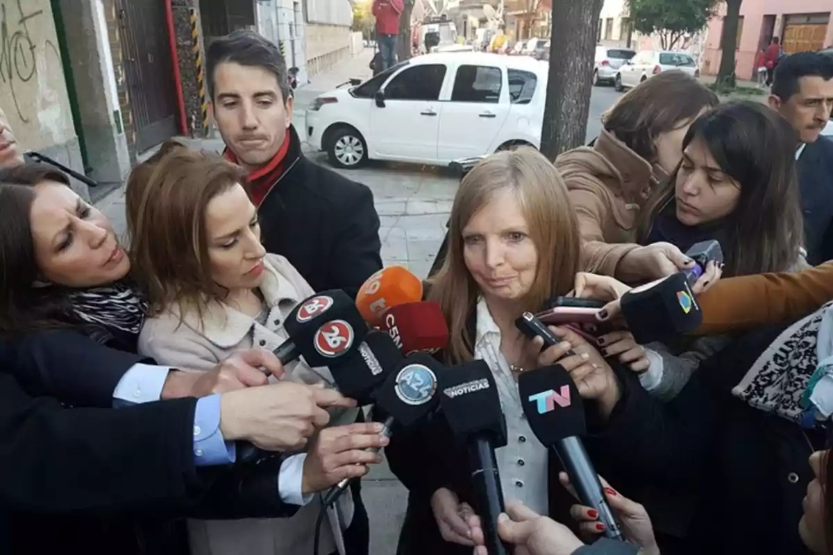 A woman surrounded by journalists and microphones on a street while answering questions A woman surrounded by journalists and microphones on a street while answering questions