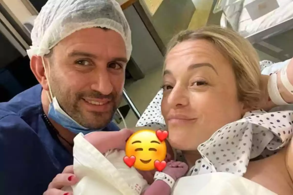 Smiling couple in a hospital room next to a newborn whose face is covered with a smiling face with hearts emoji