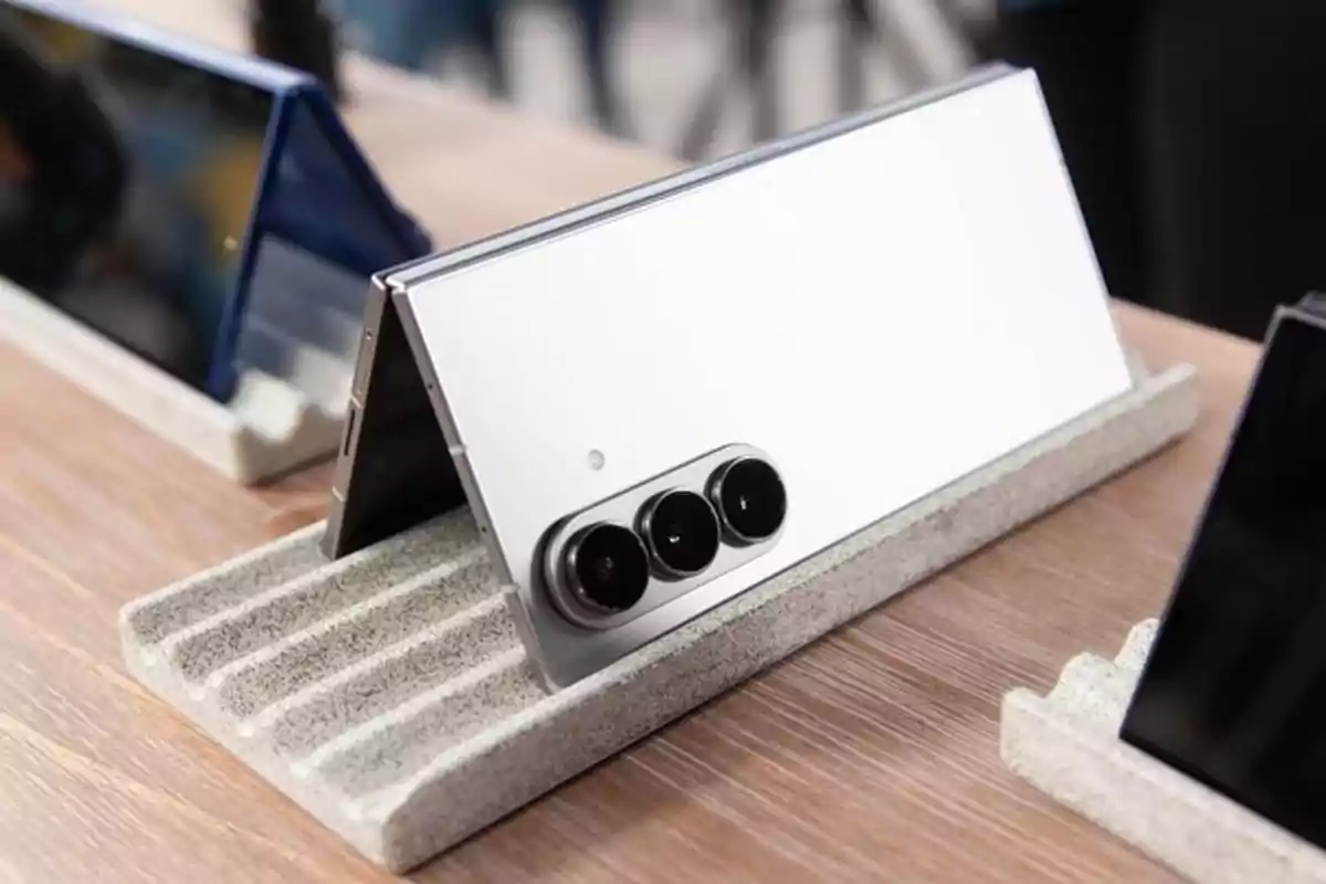 Teléfono plegable de color plateado con triple cámara trasera expuesto sobre una base en una mesa de madera Teléfono plegable de color plateado con triple cámara trasera expuesto sobre una base en una mesa de madera