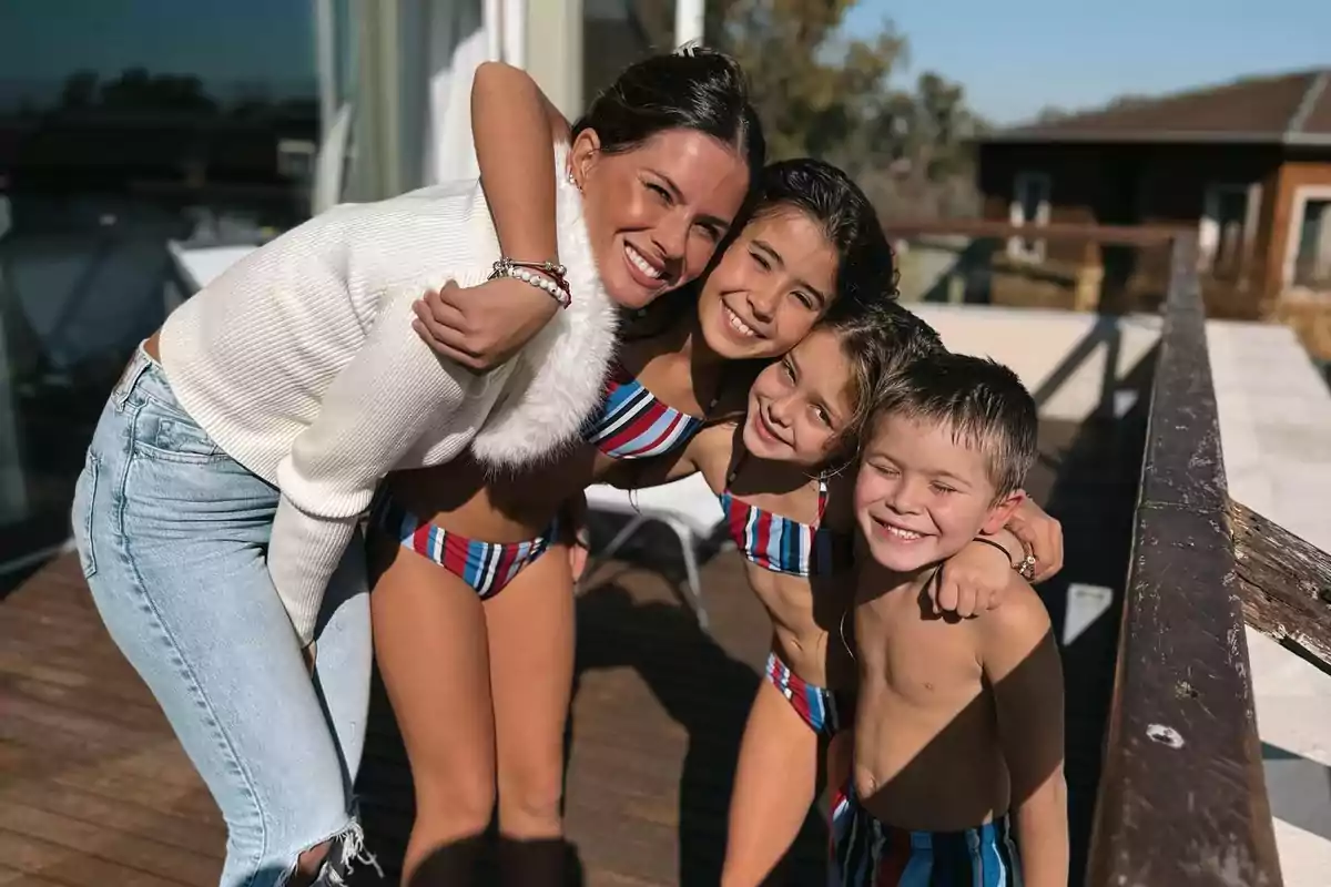 Cuatro personas sonríen abrazadas en una terraza soleada, dos niñas y un niño llevan trajes de baño a rayas y una mujer viste suéter blanco y jeans claros.