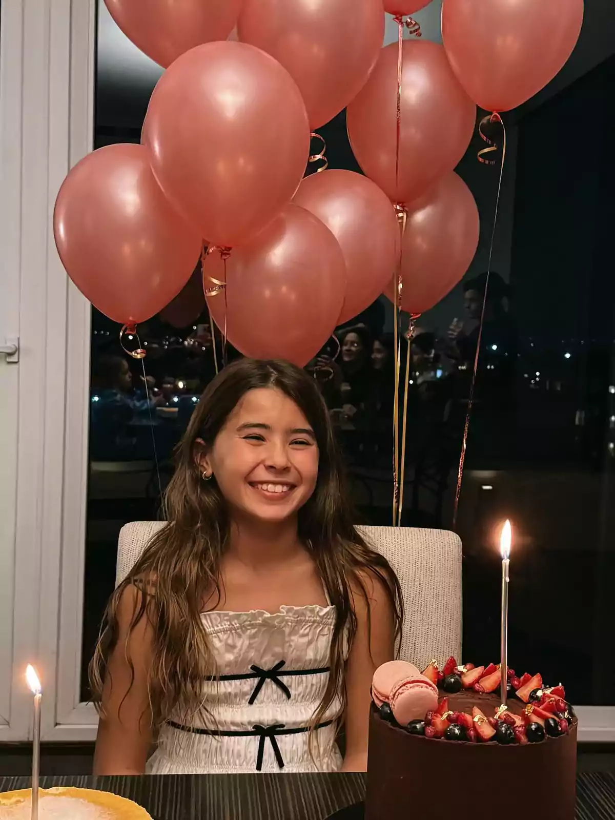 Niña sonriente sentada frente a un pastel de cumpleaños de chocolate decorado con frutas y macarons, rodeada de globos rosas en una celebración nocturna