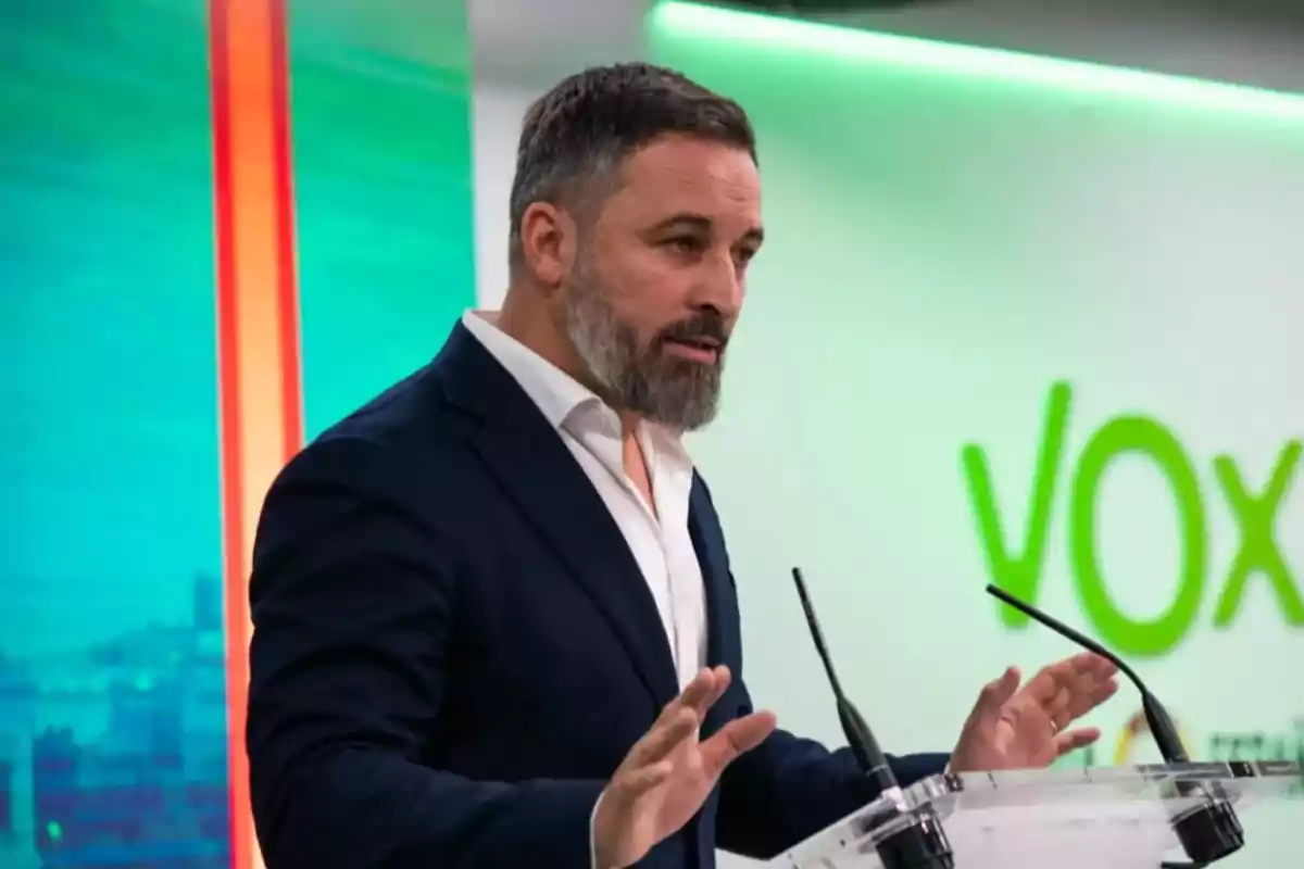 Hombre de barba y cabello corto hablando en un atril con micrófonos frente a un fondo verde con el logo de Vox Hombre de barba y cabello corto hablando en un atril con micrófonos frente a un fondo verde con el logo de Vox