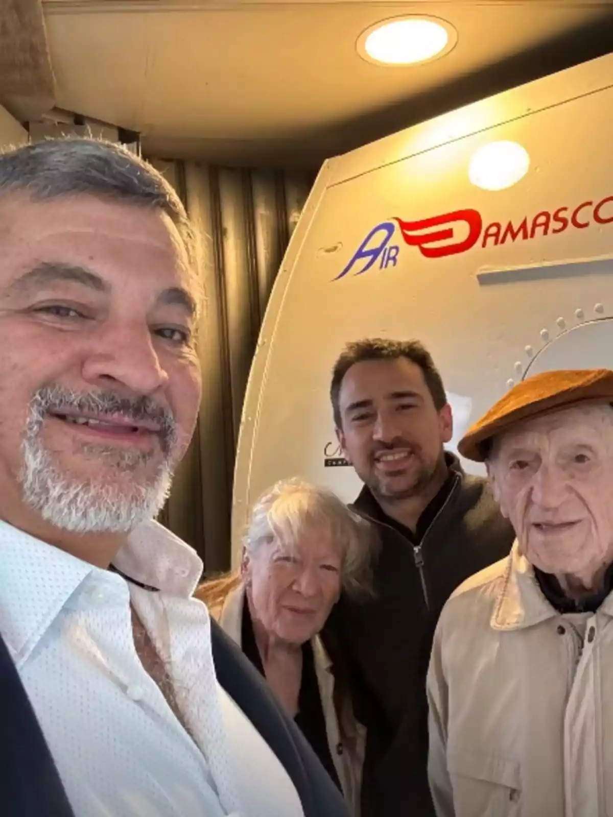 Four people smile for a selfie in front of the entrance to an Air Damascus airline plane. Four people smile for a selfie in front of the entrance to an Air Damascus airline plane.