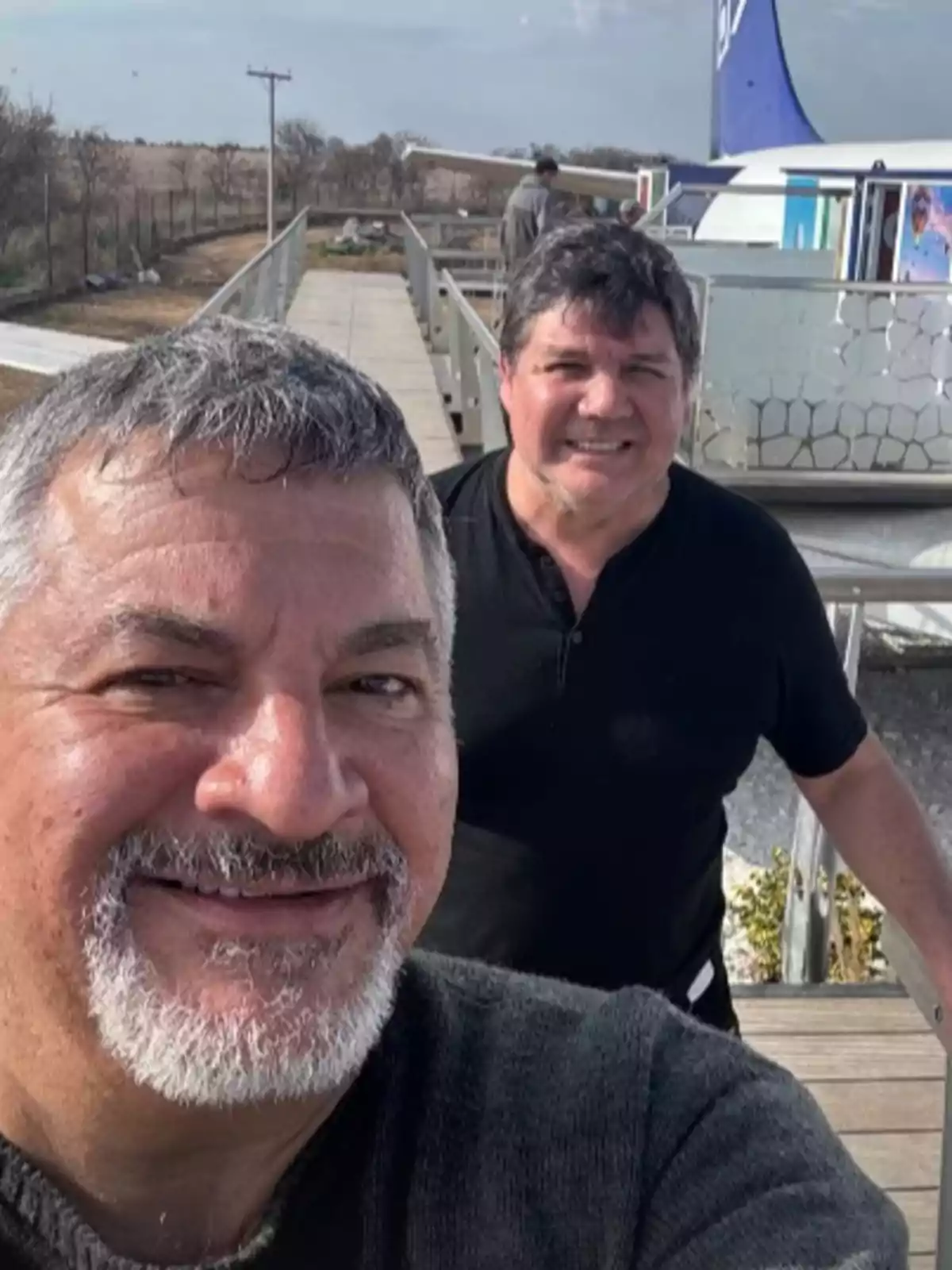 Two men smile while taking a selfie on an outdoor walkway with a plane in the background. Two men smile while taking a selfie on an outdoor walkway with a plane in the background.