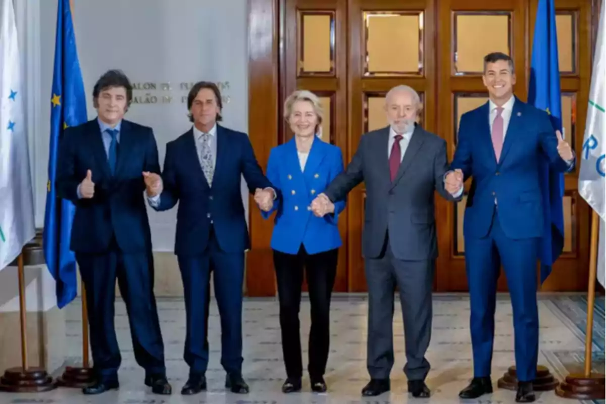 Cinco personas vestidas de traje posan de pie tomadas de las manos en un entorno formal con banderas a los lados y una puerta de madera al fondo