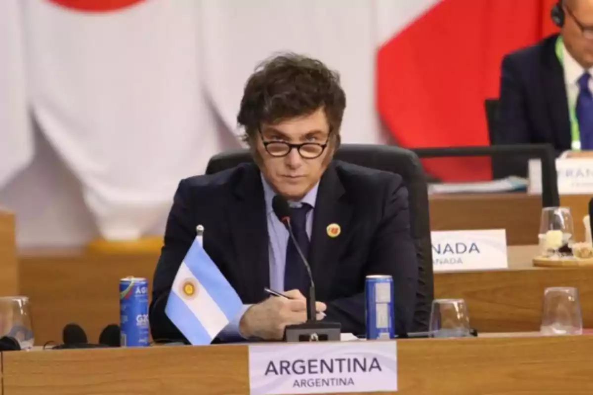 Man with glasses and a dark suit sitting at a conference table with an Argentine flag and a sign that says Argentina