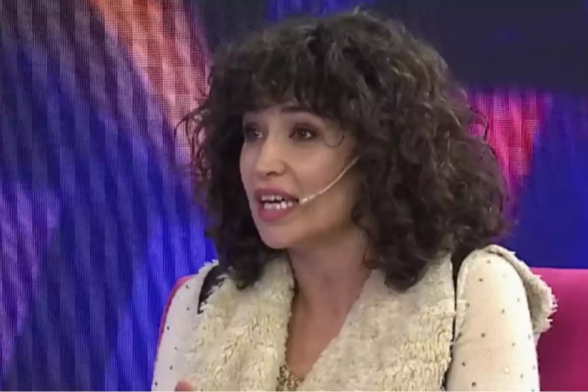 Mujer de cabello rizado y oscuro hablando en un programa de televisión con micrófono en la mejilla y fondo colorido
