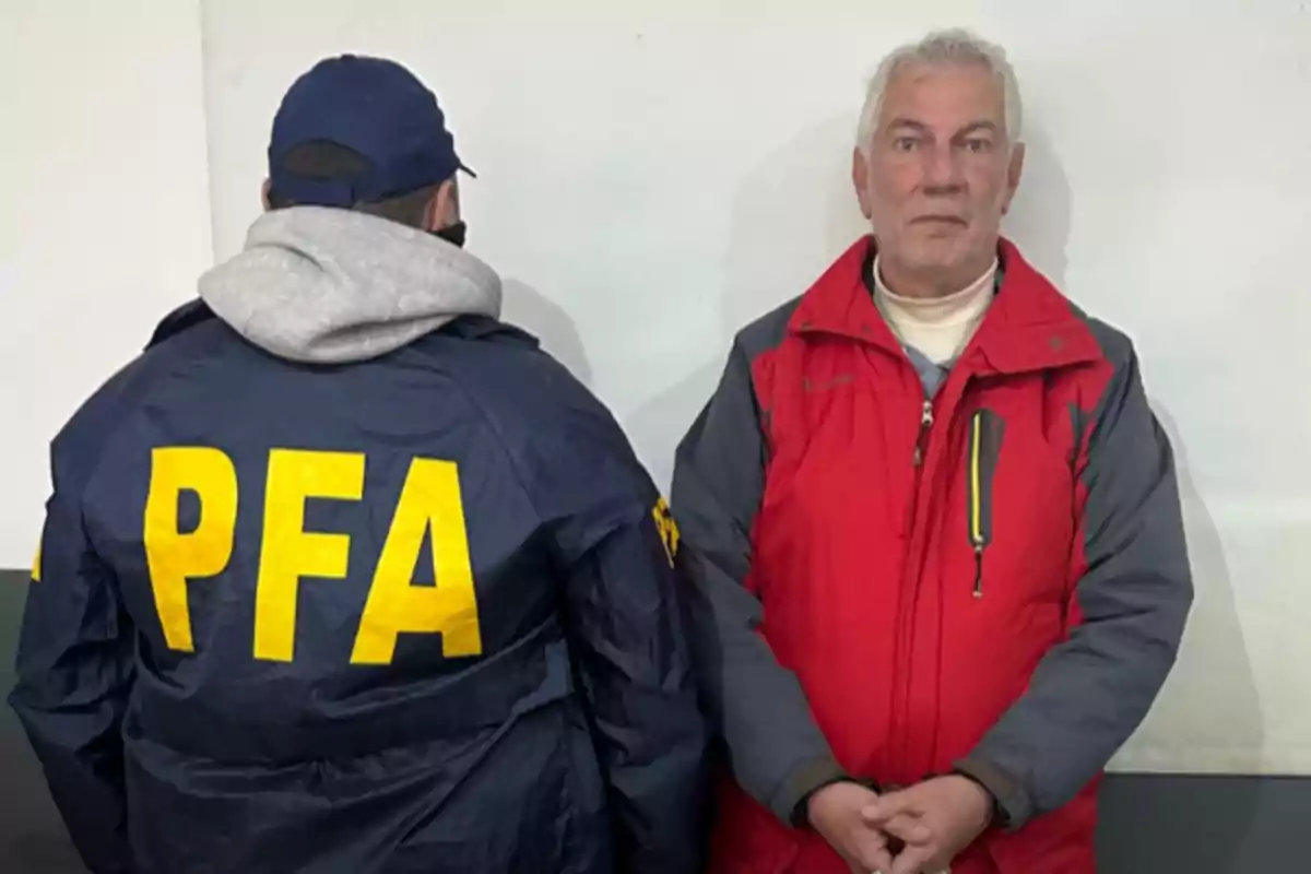 Un hombre de cabello canoso con campera roja está de pie junto a un agente de espaldas con campera azul y letras amarillas que dicen PFA frente a una pared blanca