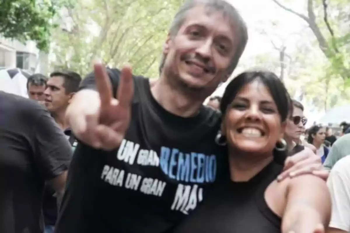 Two people smile and pose with their arms around each other at an outdoor demonstration, one of them making the victory sign with their hand.