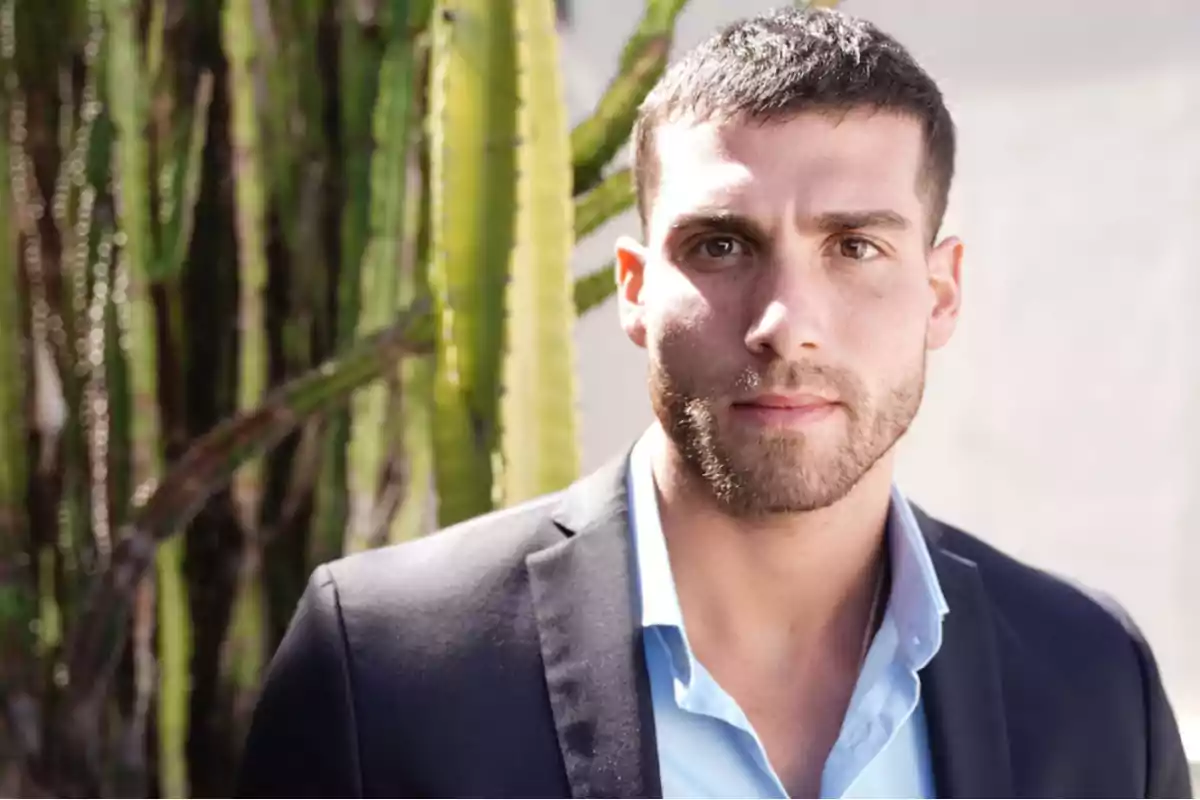 Hombre joven con barba y cabello corto viste saco oscuro y camisa azul claro posando frente a un cactus bajo la luz del sol