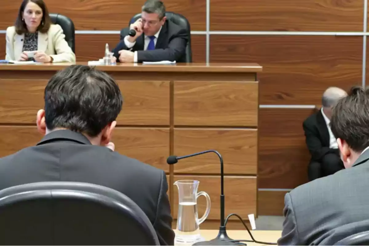 People in a courtroom with a judge and a woman seated at the front and two men seen from behind in the foreground
