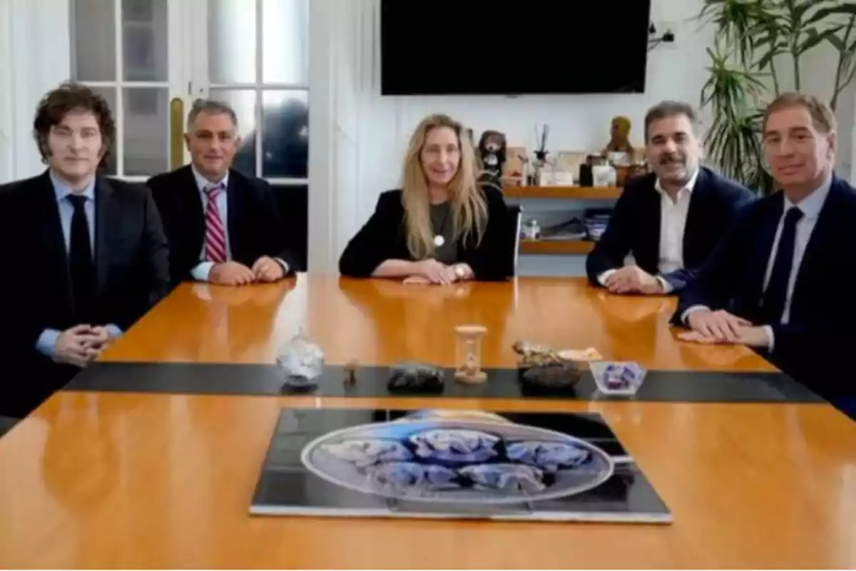 Five formally dressed people sitting around a wooden conference table with various decorative objects on the table and a large screen on the back wall.