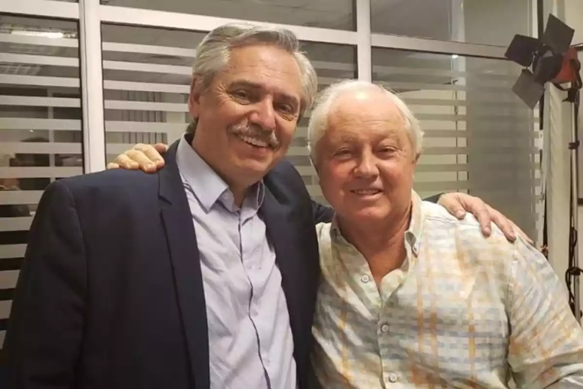 Dos hombres mayores sonríen y posan juntos en una oficina moderna con paredes de vidrio y persianas horizontales