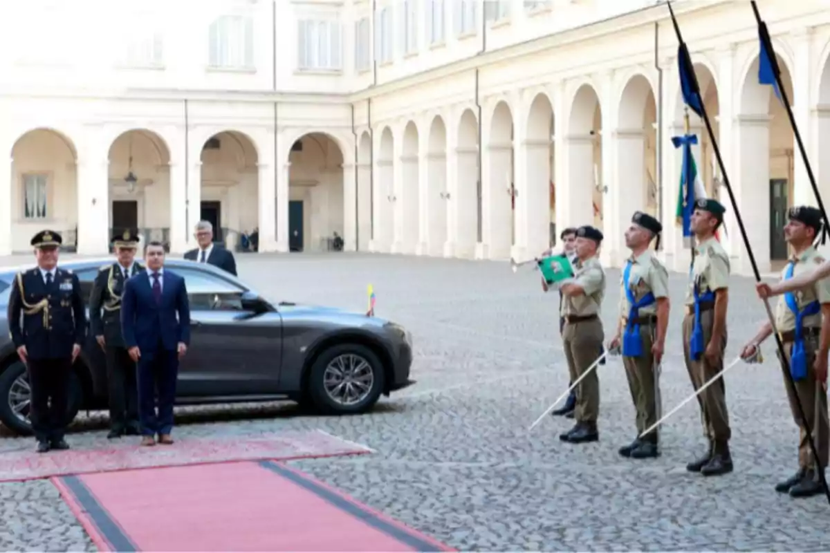 Ceremonia oficial de bienvenida con alfombra roja en un patio rodeado de arcos donde un grupo de militares uniformados sostiene banderas y espadas mientras varias personas de traje están junto a un automóvil gris Ceremonia oficial de bienvenida con alfombra roja en un patio rodeado de arcos donde un grupo de militares uniformados sostiene banderas y espadas mientras varias personas de traje están junto a un automóvil gris