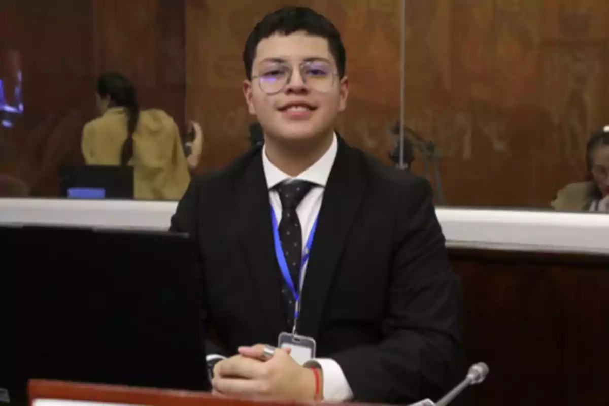 Joven con gafas y traje formal sentado frente a una computadora en una sala de conferencias