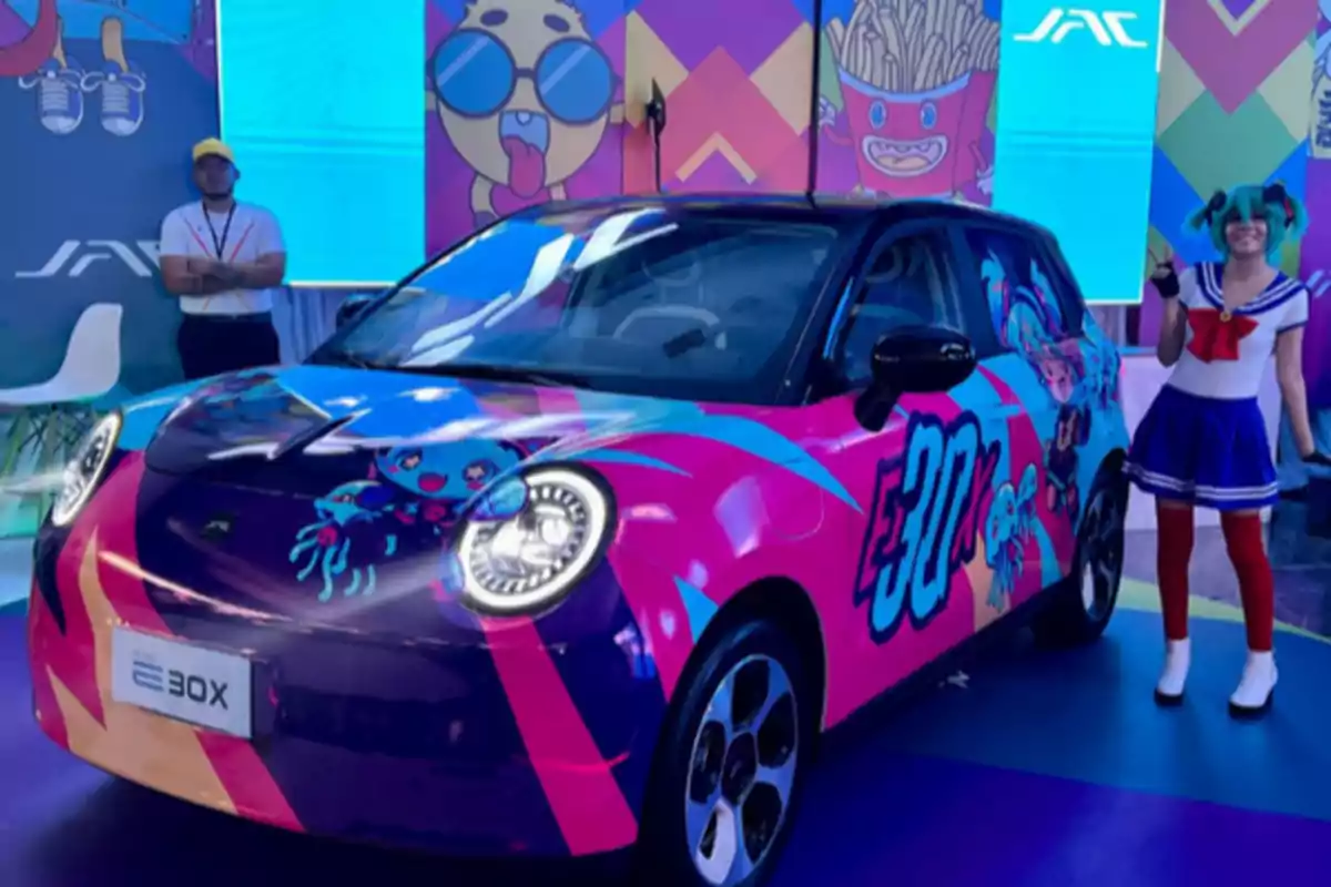 Auto eléctrico decorado con colores llamativos y personajes de estilo anime en una exhibición, junto a una persona disfrazada con un traje de marinero y otra persona de pie al fondo Auto eléctrico decorado con colores llamativos y personajes de estilo anime en una exhibición, junto a una persona disfrazada con un traje de marinero y otra persona de pie al fondo