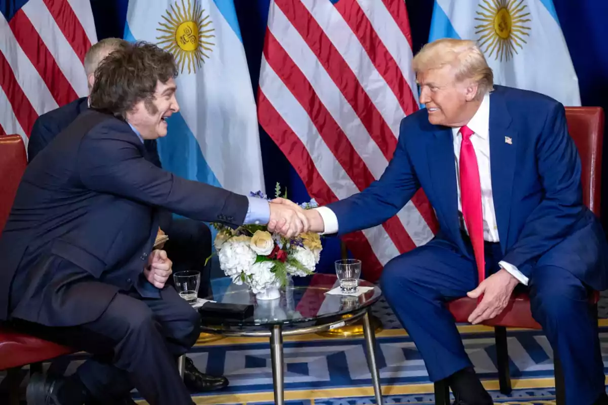 Two men in suits shake hands while sitting across a small table with the flags of Argentina and the United States in the background.