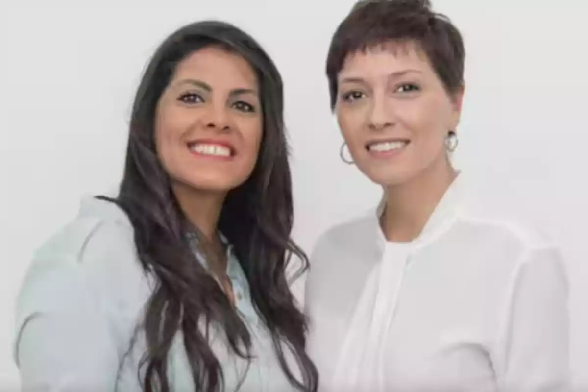 Two smiling women pose together in front of a white background. Two smiling women pose together in front of a white background.