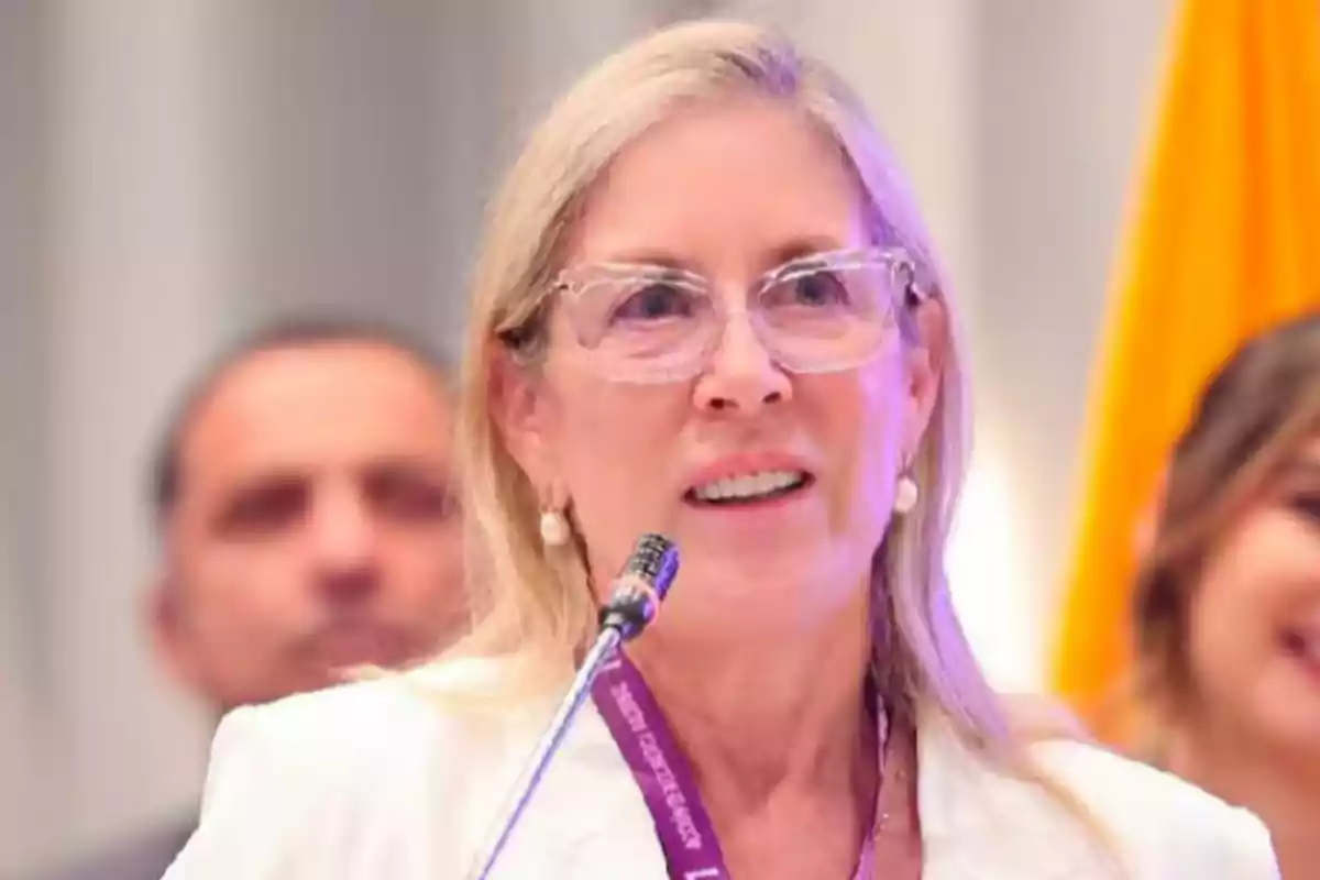 Mujer rubia con gafas y saco blanco hablando frente a un micrófono en un evento Mujer rubia con gafas y saco blanco hablando frente a un micrófono en un evento