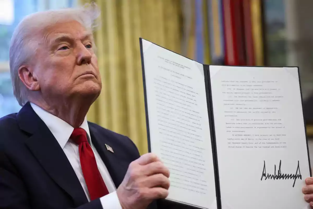 Gray-haired man in a dark suit and red tie holds an open document with text and a large signature at the bottom right. Gray-haired man in a dark suit and red tie holds an open document with text and a large signature at the bottom right.