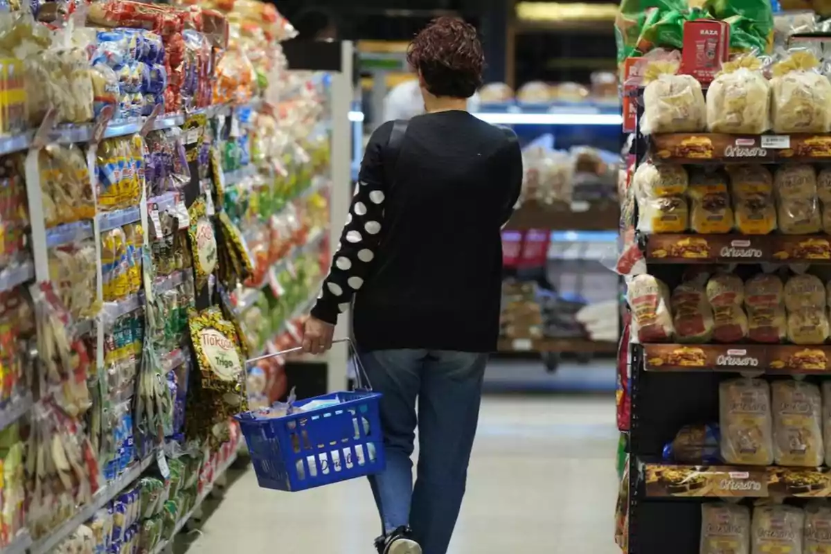 Persona de espaldas caminando por el pasillo de un supermercado con una canasta azul en la mano rodeada de estantes llenos de productos empaquetados