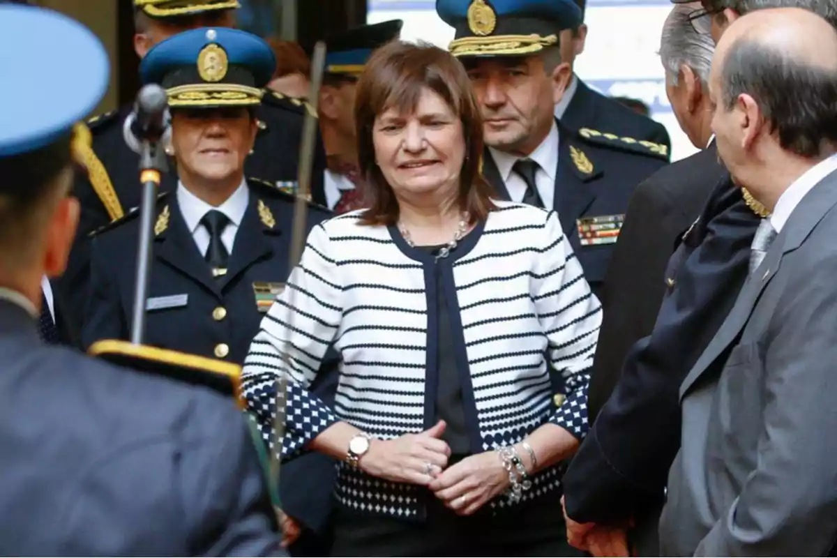 Una mujer vestida con saco blanco y negro camina entre oficiales de policía con uniformes formales y gorras azules en un evento oficial. Una mujer vestida con saco blanco y negro camina entre oficiales de policía con uniformes formales y gorras azules en un evento oficial.